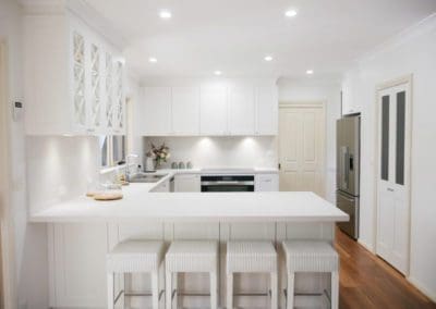 Muted white and grey Hamptons kitchen with elegant bar seating and bright kitchen ceiling lights for a warm, inviting atmosphere.