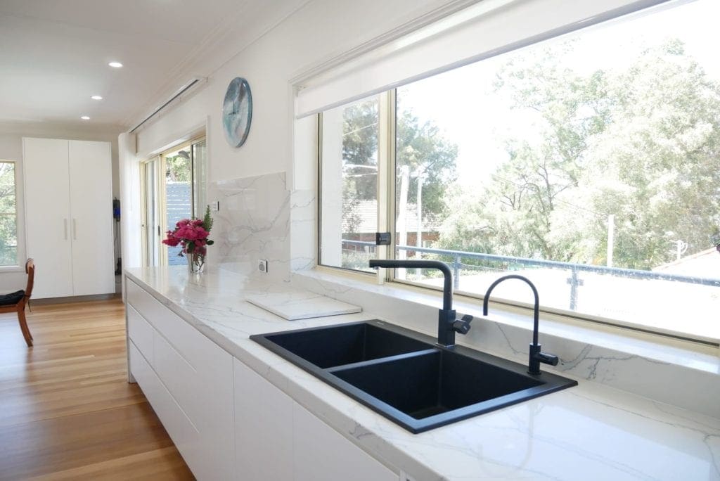 Minimalist kitchen design with white cabinetry, black dual sink, marble-look benchtops, and large windows overlooking a leafy suburban view.