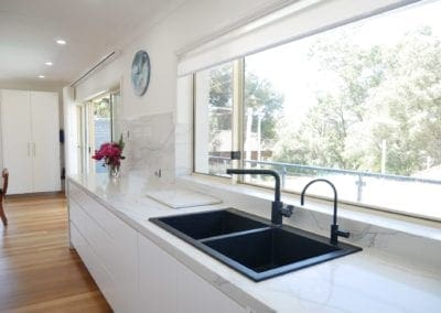 Minimalist kitchen design with white cabinetry, black dual sink, marble-look benchtops, and large windows overlooking a leafy suburban view.