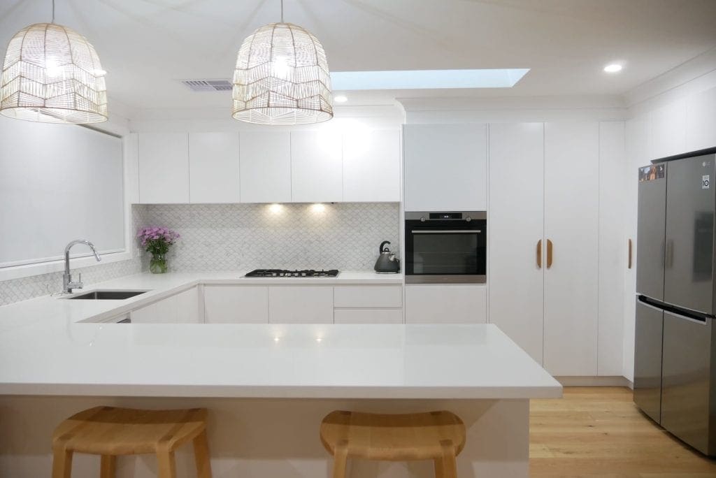 Minimalist white kitchen with a peninsula-style island bench, natural timber stools, and rattan pendant lights.