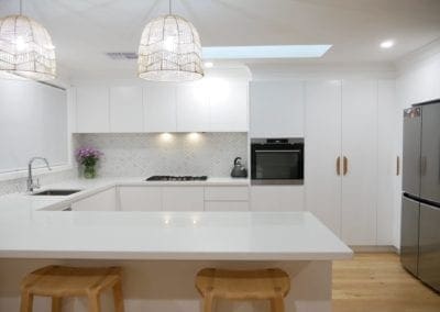 Minimalist white kitchen with a peninsula-style island bench, natural timber stools, and rattan pendant lights.