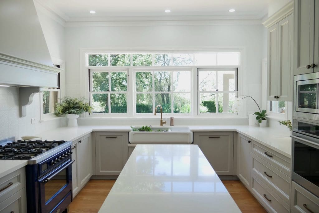 Hamptons-style kitchen featuring shaker cabinetry, farmhouse sink, brushed brass tapware, and a large window overlooking a lush green garden.