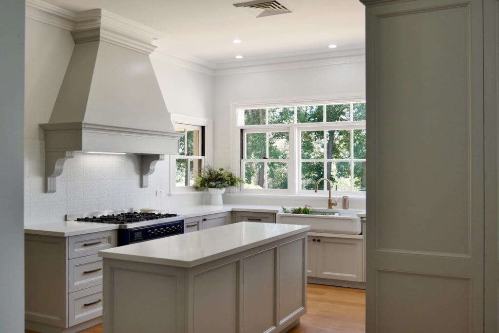 Modern farmhouse kitchen with a central island, shaker-style cabinetry, elegant range hood, farmhouse sink, and large windows offering a view of lush greenery.