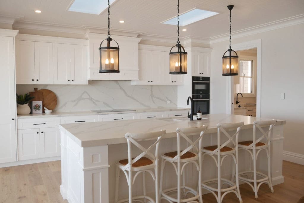 A luxurious ocean-inspired kitchen featuring sleek white Dulux Vivid White polyurethane cabinetry, Smartstone Borghini Naturale benchtops, and integrated Bosch appliances. The kitchen is designed to complement a stunning coastal view, with elegant black accents on the faucet and hardware, and a spacious island with a clean, modern aesthetic. The design flows seamlessly into a covered balcony for effortless entertaining.