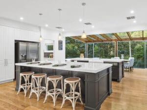 Modern open-plan kitchen design featuring a large island with dark cabinetry, white stone benchtops, natural timber floors, and floor-to-ceiling glass windows overlooking a lush garden.