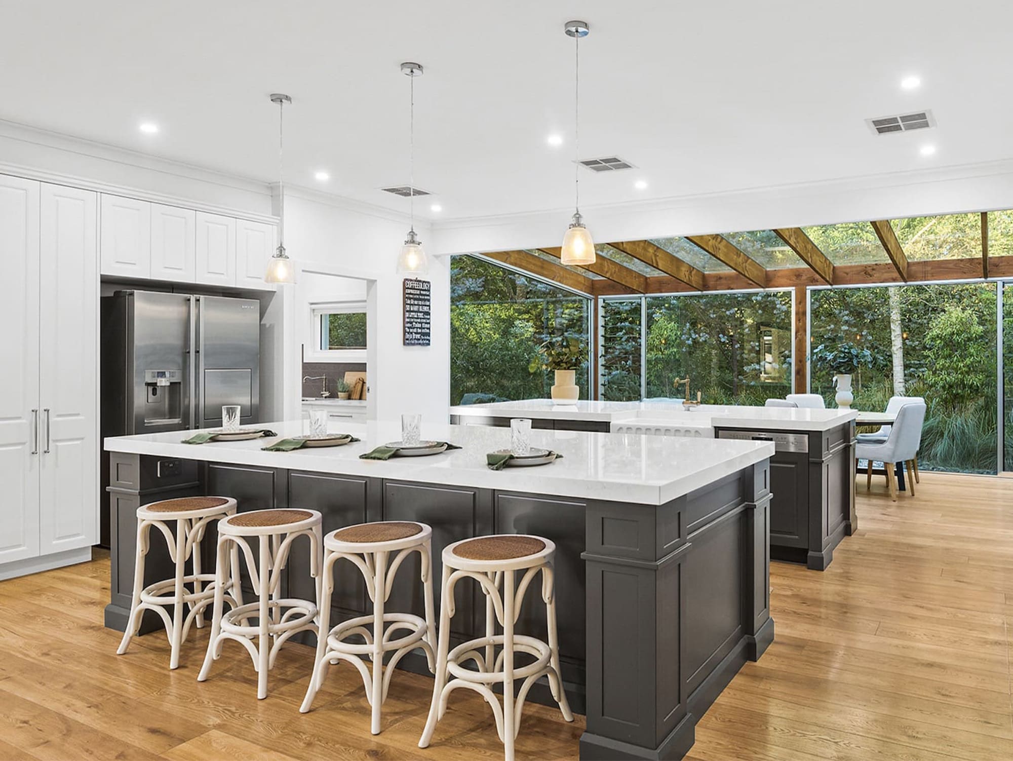 Modern open-plan kitchen design featuring a large island with dark cabinetry, white stone benchtops, natural timber floors, and floor-to-ceiling glass windows overlooking a lush garden.
