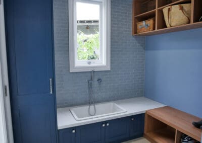 The laundry/mudroom features colour-matched blue cabinetry with dark anthracite carcasses. The benchtop continues the kitchen theme with YDL Angel Falls, while the box shelves are made from Polytec Tasmanian Oak Woodmatt. A Cuisine drop-in dog bath is conveniently placed at floor level, and a Cuisine sink sits under the window, framed by hand-laid splashback matching tiles.