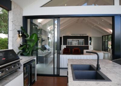 The alfresco kitchen features benchtops and an island encased in Tundra Grey limestone, complementing the rustic wall cladding on the exterior columns.