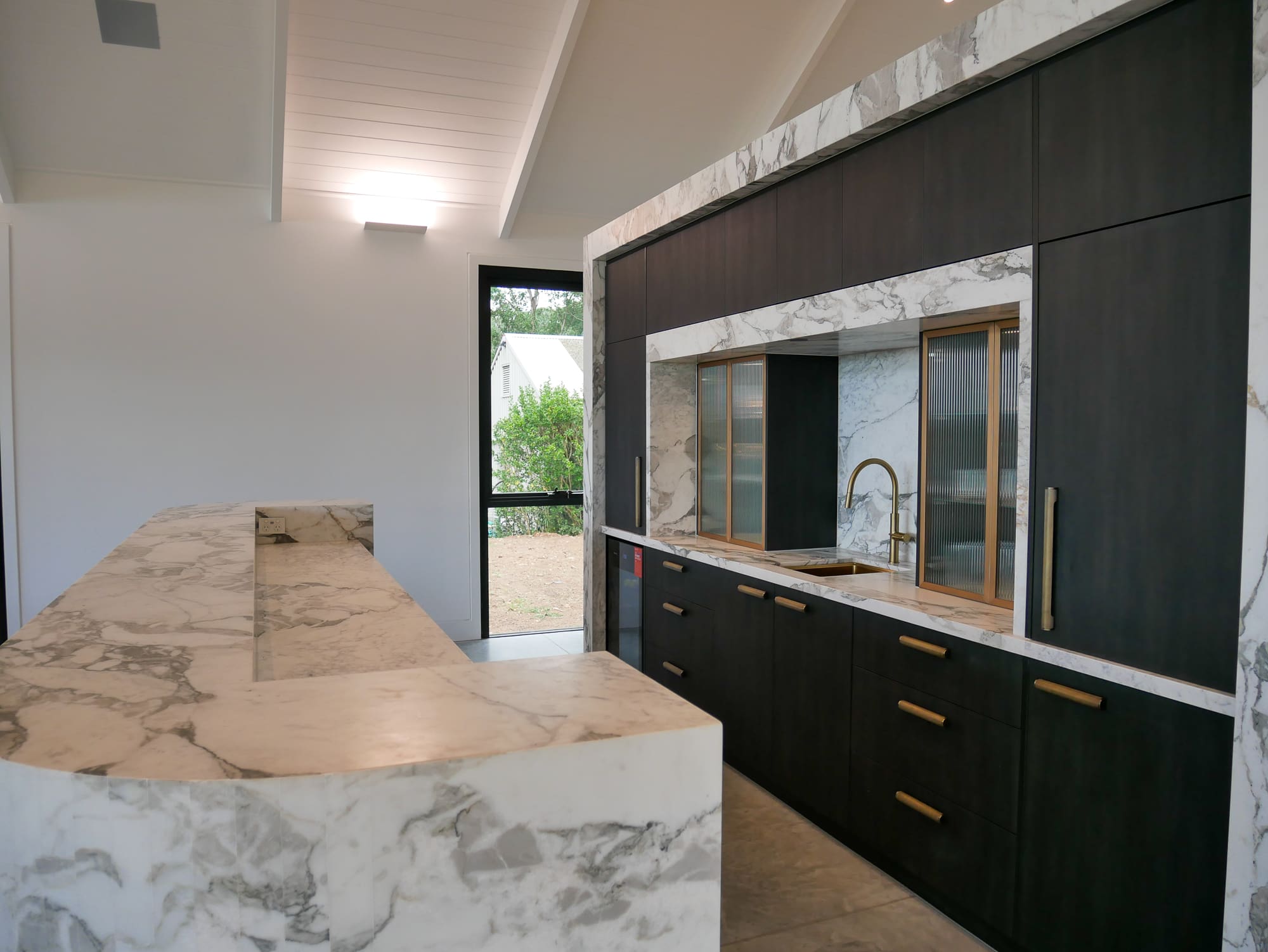 Bar side of the kitchen featuring a full Calacatta Vagili marble splashback, Titan brass undermount sink, and matching brass Titan tap. Aluminium doors with brass finish and fluted glass inserts frame the bar, with glass shelves inside the push-to-open doors.