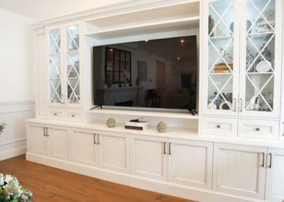 Matching hand-painted TV unit and display cabinet in the living room with X-framed glass panels, complementing the kitchen's pendants and showcasing treasured items with internal upper cabinetry lighting.