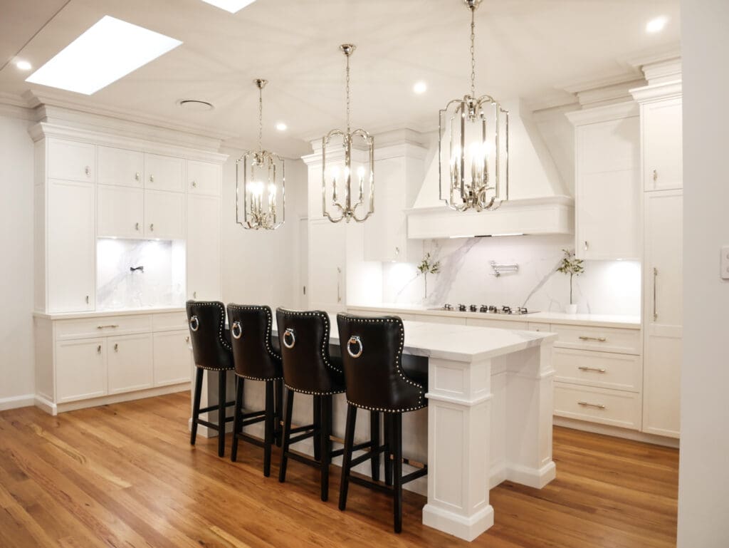 Kitchen featuring three polished nickel pendants over a stunning island with leather bar chairs. Dulux Lexicon quarter strength matte finish and modern Colorado profile cabinetry with decorative capping. Quantum Quartz Alpine White benchtops and Classico Palazzo splashbacks with grey veining, complementing the elegant design.