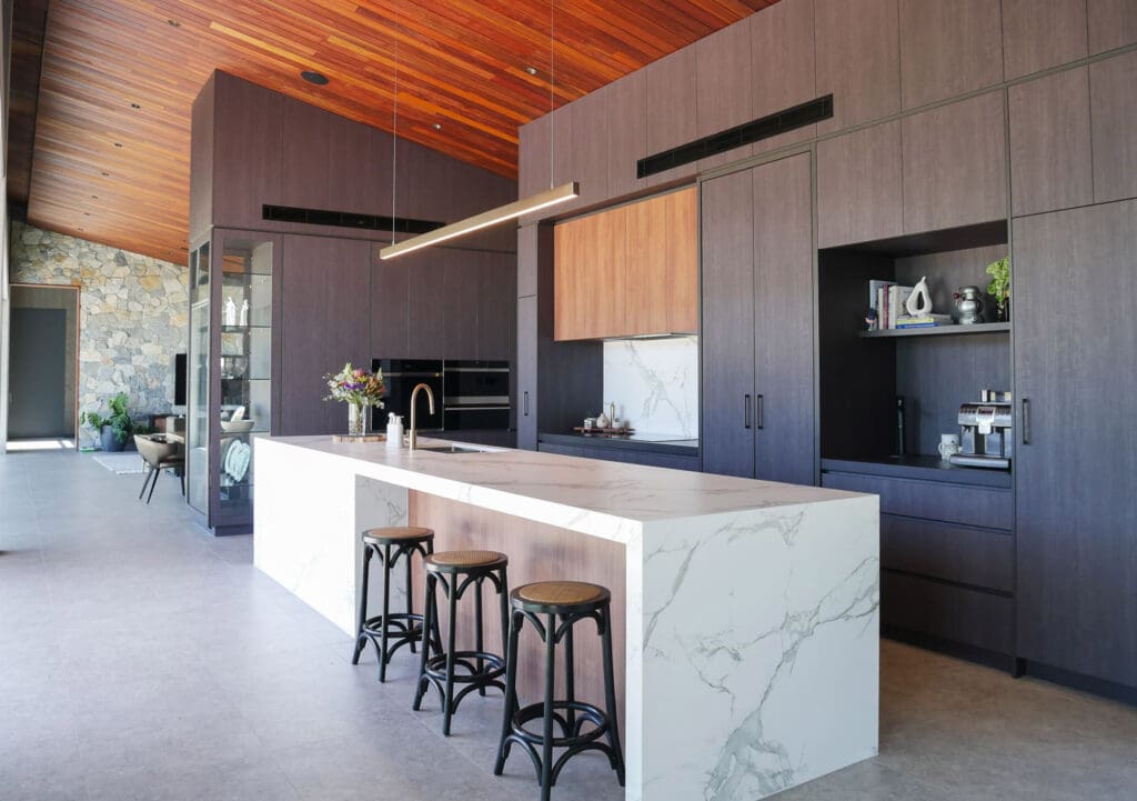 Side view of a two-toned kitchen with soaring ceilings, showcasing Dekton Sirius porcelain benchtops for the sink area and Atlas Plan Statuario Supremo Silk with white and grey veins for the island and splashback. The 60mm mitred edges and 80mm island edges complement the grey concrete-look flooring.