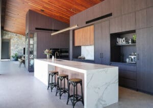 Side view of a two-toned kitchen with soaring ceilings, showcasing Dekton Sirius porcelain benchtops for the sink area and Atlas Plan Statuario Supremo Silk with white and grey veins for the island and splashback. The 60mm mitred edges and 80mm island edges complement the grey concrete-look flooring.