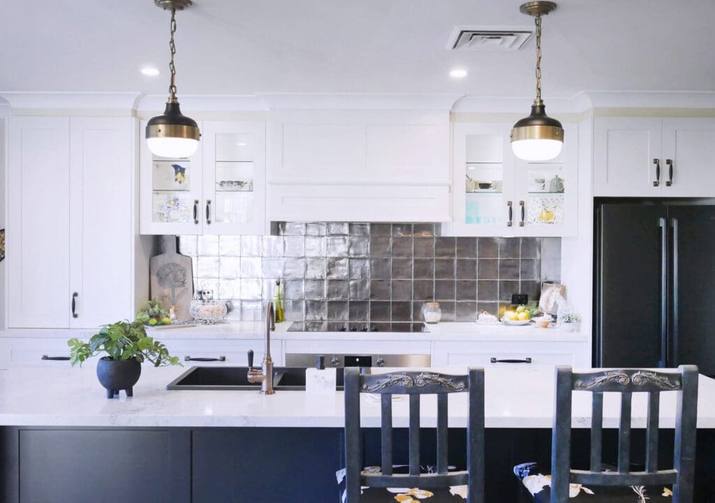 Brushed brass and matte black pendants over the island complement the Statue Bronze Brodware Winslow tap, black granite sink, and handmade brass and black wooden handles. The Bosch rangehood with glass inset doors and underbench lighting highlights a unique Pewter-finished splashback with handmade Spanish tiles.
