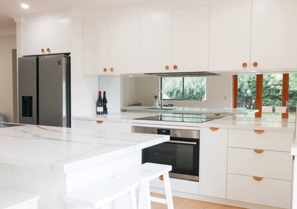 Kethy Olympia Oak knobs on doors and L7804 lip handles on drawers complement the natural oak floors and benchtop. The 40mm thick Calacatta Oro from Smartstone features sweeping veins and a waterfall edge, while the mirrored glass splashback reflects garden greenery, enhancing the open plan design.