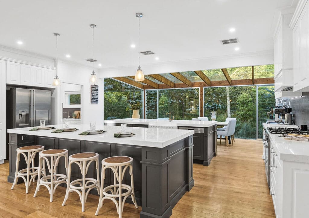 Spacious modern kitchen with a large central island, pendant lighting, and floor-to-ceiling glass windows overlooking lush greenery.