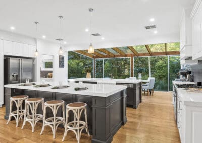 Spacious modern kitchen with a large central island, pendant lighting, and floor-to-ceiling glass windows overlooking lush greenery.