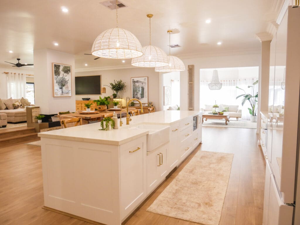 Spacious, light-filled kitchen with a large white island featuring gold fixtures, farmhouse sink, and pendant rattan lighting, opening into a cozy, modern living area with wooden floors and natural decor.