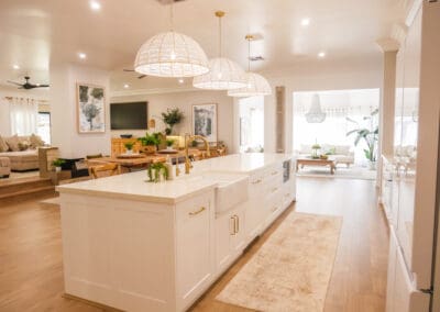 Spacious, light-filled kitchen with a large white island featuring gold fixtures, farmhouse sink, and pendant rattan lighting, opening into a cozy, modern living area with wooden floors and natural decor.