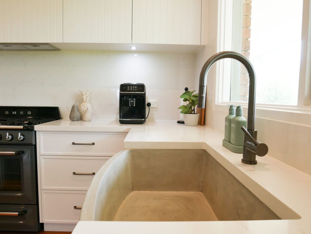 Close-up of a modern farmhouse kitchen sink with a deep concrete basin, brass pull-down faucet, white stone countertops, and shaker-style white cabinetry. A Smeg oven, coffee machine, and minimal decorative items complete the stylish setup.