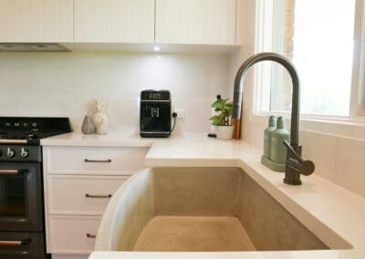 Close-up of a modern farmhouse kitchen sink with a deep concrete basin, brass pull-down faucet, white stone countertops, and shaker-style white cabinetry. A Smeg oven, coffee machine, and minimal decorative items complete the stylish setup.