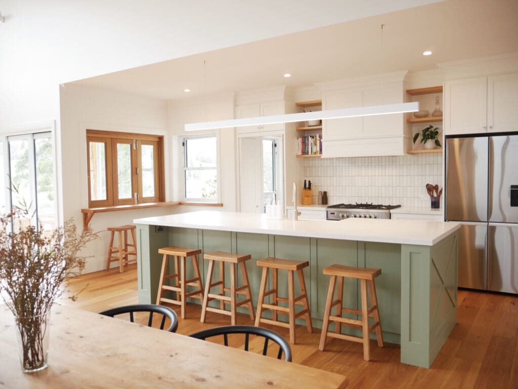 Modern farmhouse kitchen featuring a large island with sage green paneling, wooden bar stools, white cabinetry, stainless steel appliances, open shelving with decor and books, and warm timber flooring.