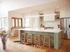 Modern farmhouse kitchen featuring a large island with sage green paneling, wooden bar stools, white cabinetry, stainless steel appliances, open shelving with decor and books, and warm timber flooring.