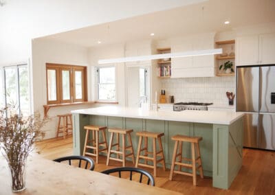 Modern farmhouse kitchen featuring a large island with sage green paneling, wooden bar stools, white cabinetry, stainless steel appliances, open shelving with decor and books, and warm timber flooring.