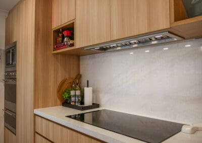 Stainless steel appliances, including a Smeg oven, Smeg cooktop, and Whispair rangehood, are integrated into the wall cabinetry, creating a visual break and complementing the natural wood patterns in the kitchen design.