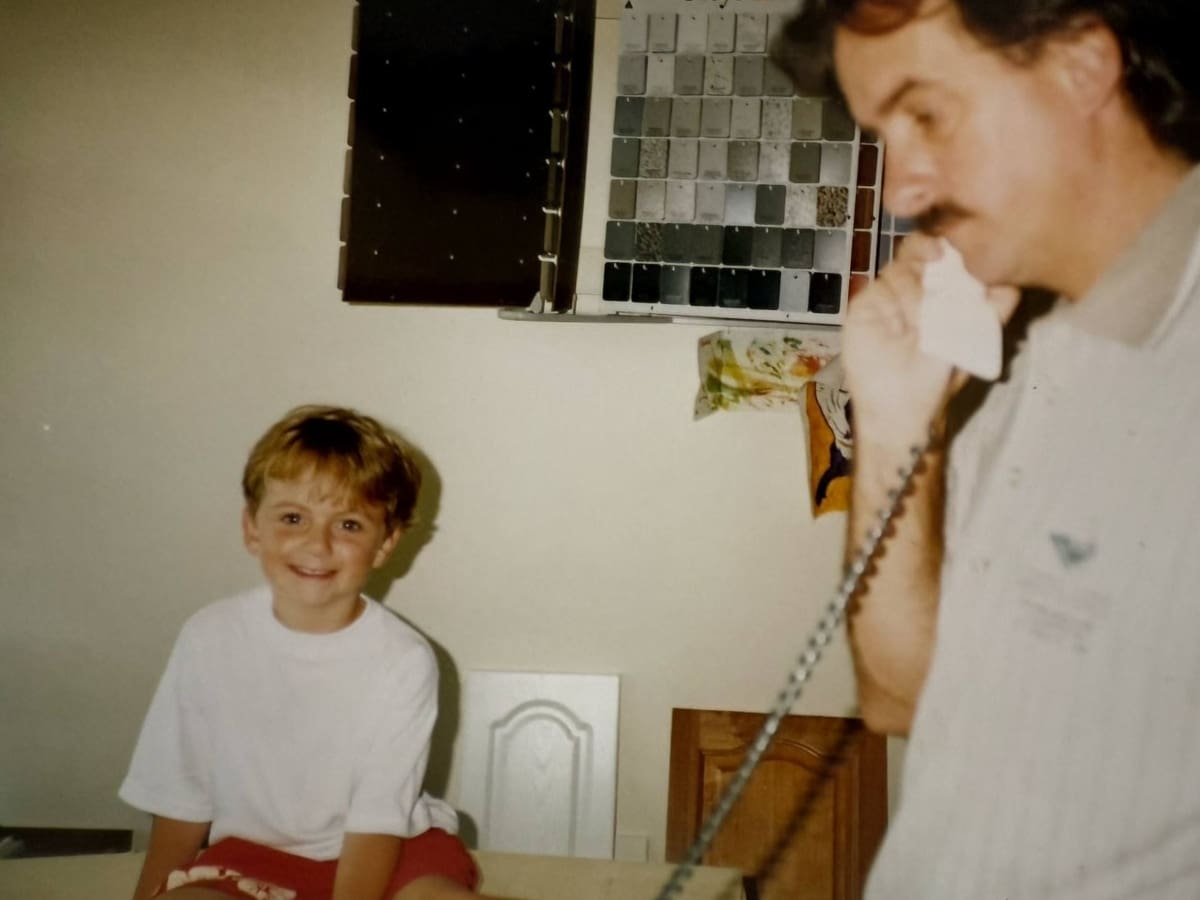 A nostalgic photo of a young James smiling while seated on a kitchen counter, with Mark Clark, founder of Harrington Kitchens, in the background speaking on a corded phone. Behind them are cabinet samples — a glimpse into the early days of Harrington Kitchens and its family-run beginnings.