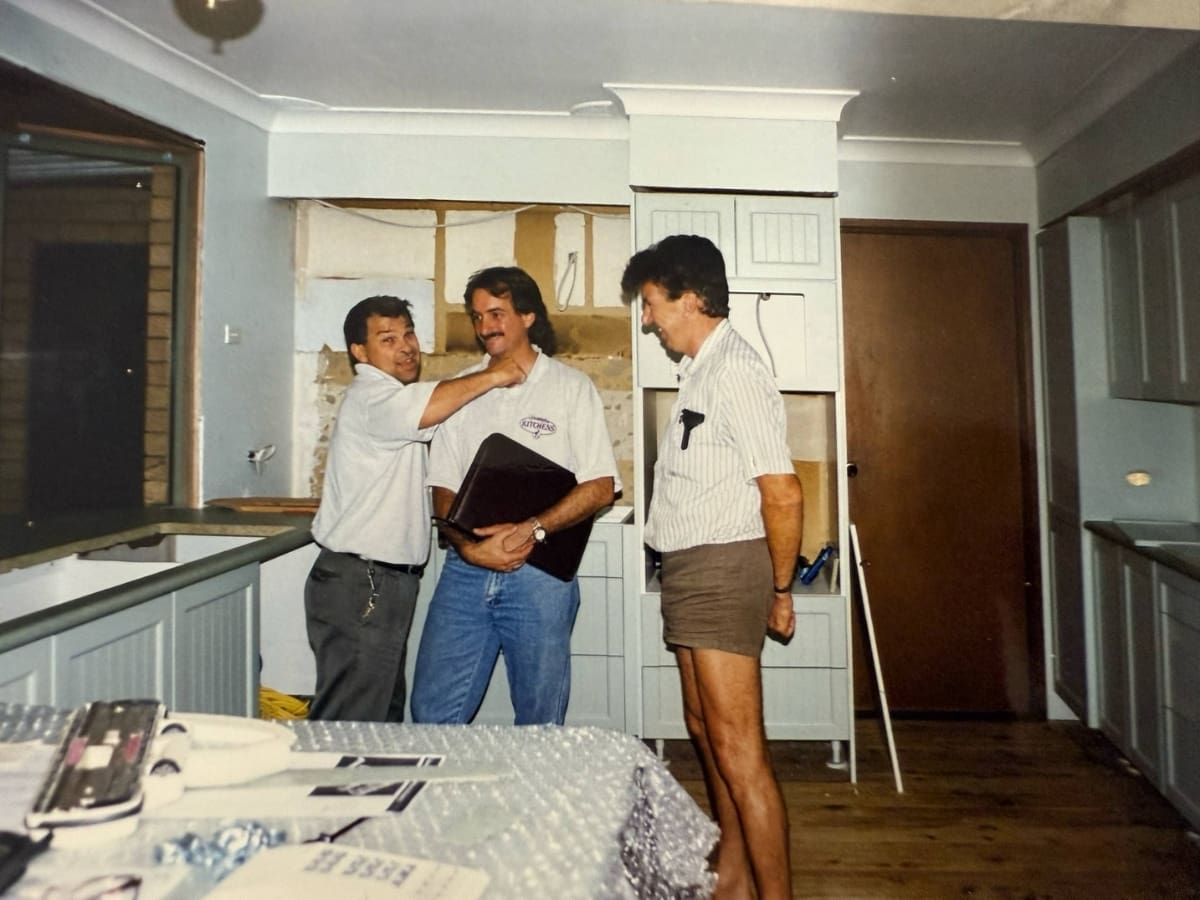 A candid moment during the early days of Harrington Kitchens — three men, including founder Mark Clark (center), share a laugh while standing in a partially completed kitchen. The space is mid-renovation, with cabinetry being installed, design plans on the table, and tools nearby — capturing the hands-on spirit and camaraderie that built the Harrington Kitchens legacy.