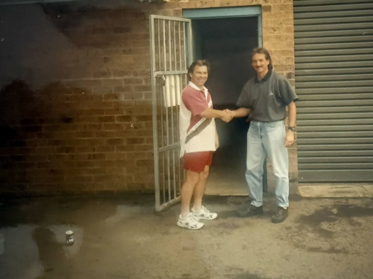 Mark Clark and a colleague shaking hands outside the first Harrington Kitchens factory in Narellan — a milestone moment marking the beginning of the company’s workshop journey. The brick building, roller door, and open gate reflect the humble yet ambitious early days.