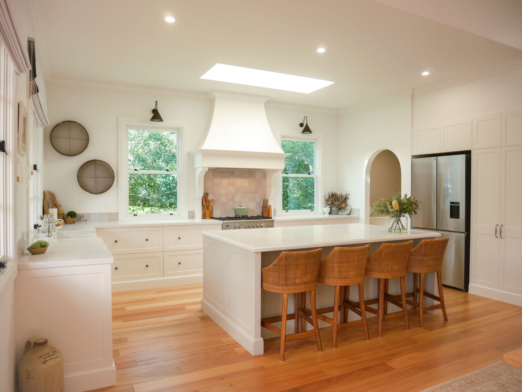 Refined Country Kitchen with Natural Light and Timeless Elegance Refined country kitchen with white shaker cabinets, central island with bespoke curved rattan stools , timber flooring, large range hood, and two windows bringing in natural light.