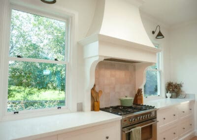 Country kitchen with custom white range hood, stainless steel oven, and shaker cabinets under two large windows.