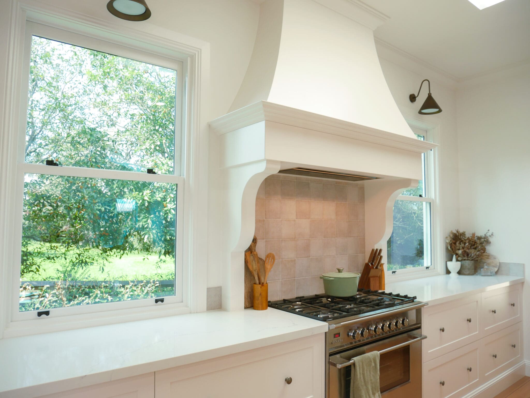 Classic Country Kitchen with Custom Range Hood and Dual Windows Country kitchen with custom white range hood, stainless steel oven, and shaker cabinets under two large windows.