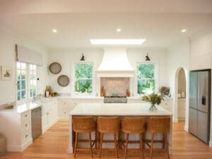 Light-filled refined country kitchen with a central island, woven bar stools, white cabinetry, timber flooring, and greenery visible through double windows.