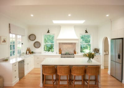 Light-filled refined country kitchen with a central island, woven bar stools, white cabinetry, timber flooring, and greenery visible through double windows.