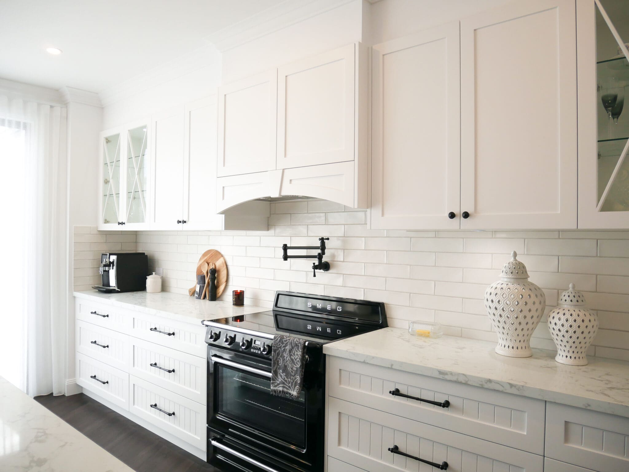 White Modern Provincial Kitchen Custom-built white rangehood above a black SMEG oven with matte black pot filler and subway tile splashback.