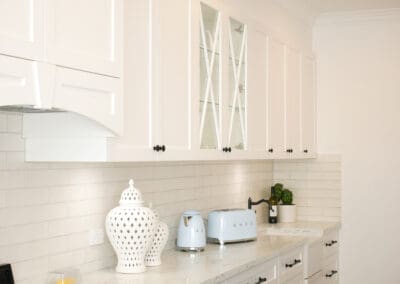 White shaker-style kitchen cabinets with glass-front upper cupboards, marble-look benchtop, and soft neutral backsplash.