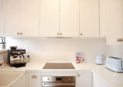 Bright white kitchen with shaker-style cabinetry, marble-look benchtops, and modern appliances including a black espresso machine and pastel blue SMEG kettle and toaster.
