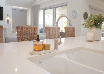 A brushed nickel Phoenix Cromford mixer tap installed over a white double 1901 Butler sink, set into a stone island benchtop. In the background, wicker bar stools line the island, and a neutral open-plan living space with large windows and natural light is visible.