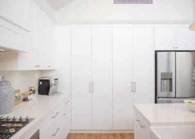 A white L-shaped kitchen with classic shaker cabinetry, tall storage cabinets surrounding a stainless steel fridge, and a seamless layout that maximises space and style.