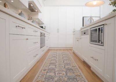 A bright L-shaped-style kitchen featuring white shaker-style cabinets, integrated stainless steel appliances, a farmhouse sink, and a patterned runner rug over light timber flooring. The design includes a built-in microwave, ample storage, and a clean, minimalist aesthetic with modern lighting and brushed nickel hardware.