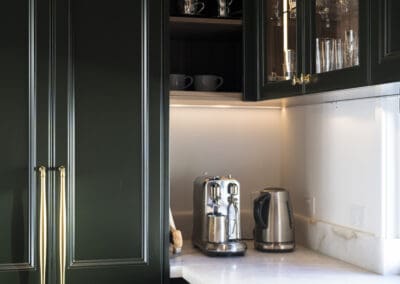 Butler’s pantry with deep green cabinetry, gold hardware, white stone benchtops, and a coffee station with espresso machine and kettle.