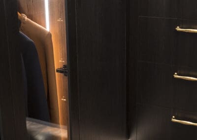 Dark timber robe with gold handles and glass panel doors, featuring soft integrated lighting and neatly hung garments.