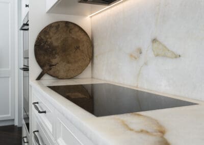 Close-up of a quartzite splashback and benchtop with integrated induction cooktop, white shaker cabinetry, and dark bronze hardware.