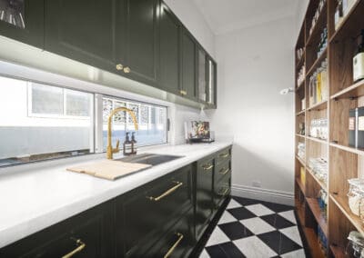 Butler’s pantry with deep green cabinetry, gold hardware, white stone benchtops, and open timber shelving.