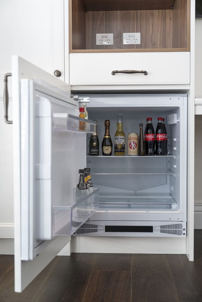 Open under-bench bar fridge with beverages, set within white custom cabinetry and timber shelving.
