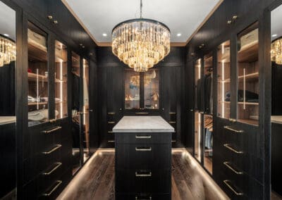 Walk-in robe with dark timber cabinetry, brass hardware, glass-fronted doors, LED lighting, central island with marble top, and a statement chandelier.