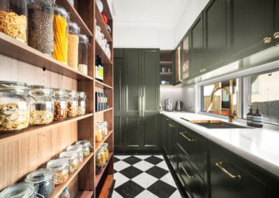 Butler’s pantry with deep green cabinetry, gold hardware, white stone benchtops, and open timber shelving.